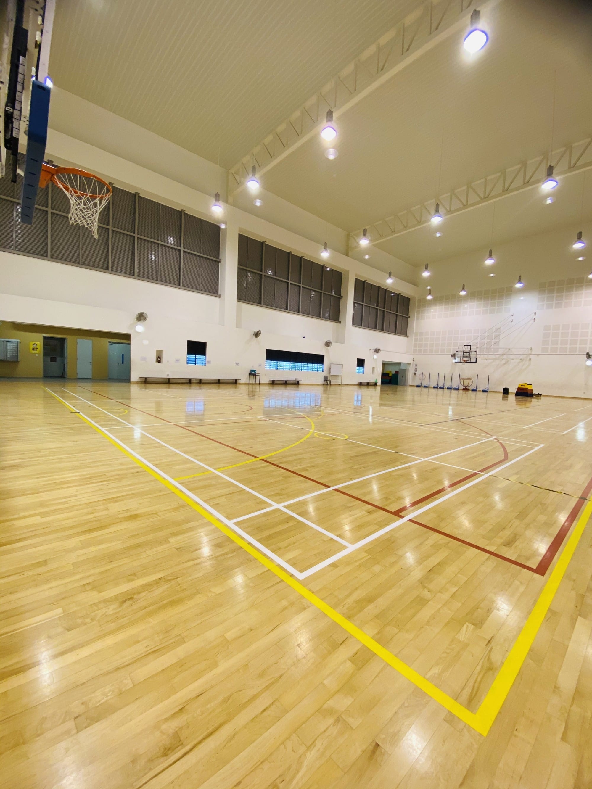 Empty indoor sports hall with a wooden floor, colored court lines, a basketball hoop, and bright overhead lights.